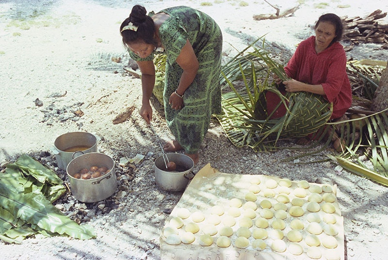 A woman prepares bread to fry in a pot, while another woman weaves with flax. They are sitting on a beach
