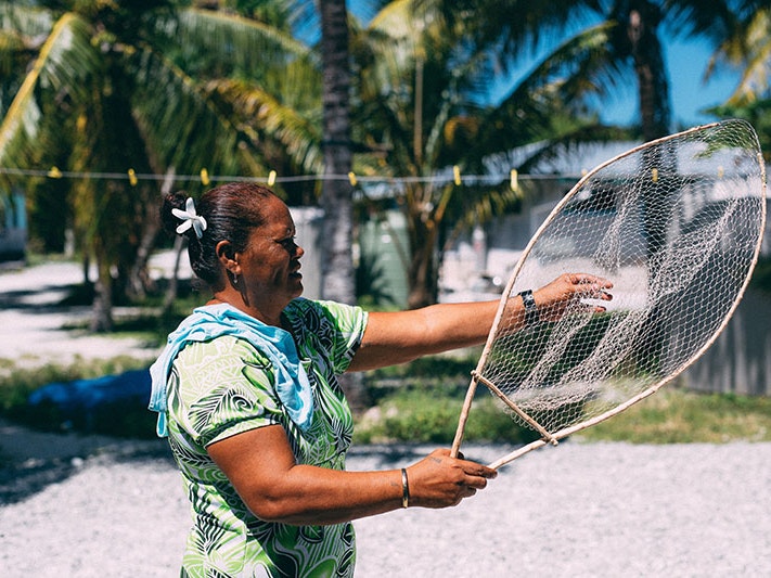 Co-collector Siniva Kalolo holding fishing net, Atafu, Tokelau, Nov 2017, Tokelau, by Andrew Matautia. Co-collected, 2017. Te Papa (O.047818) Woman holds a fishing net. Behind her is a house and palm trees