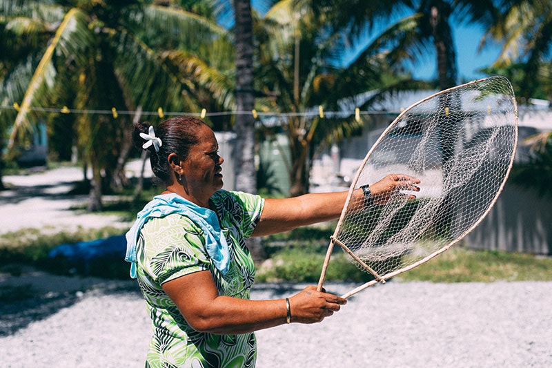 Woman holds a fishing net. Behind her is a house and palm trees