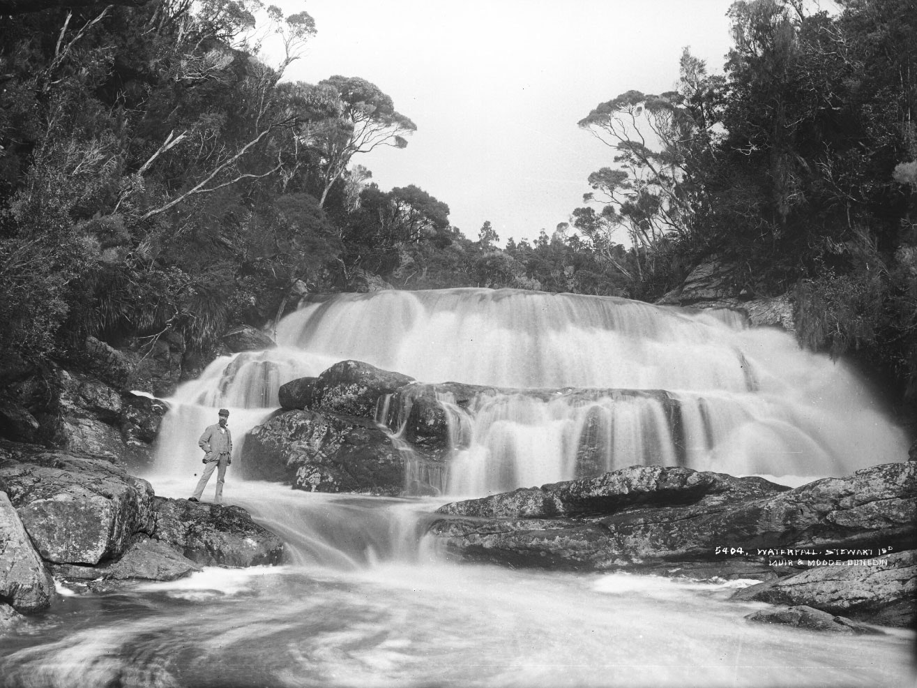 Black and white photo of a man standing in front of a large waterfall