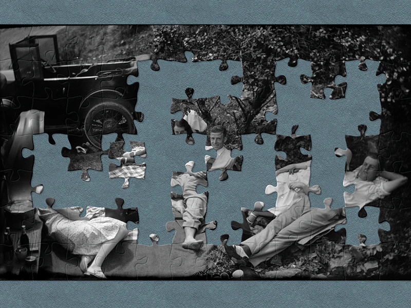 A black and white photo of five people lying on blankets having a picnic in the 1930s, they are surrounded by cars. There are jigsaw-shaped pieces missing from the image
