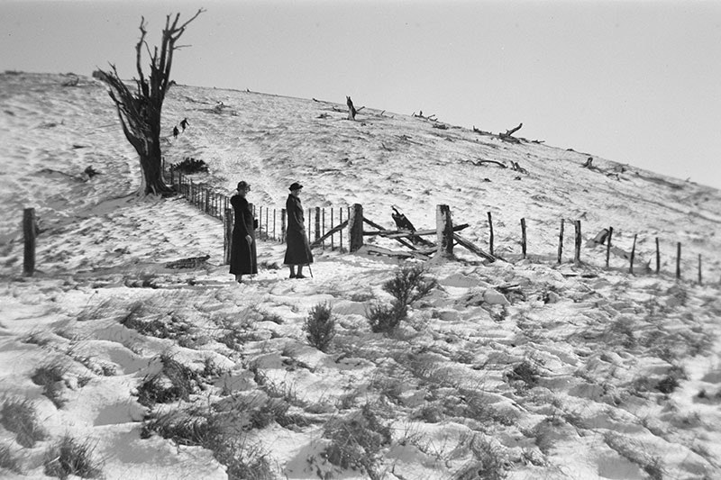 Two women in formal coats and hats stand for a photo in a field covered in snow, beside a fence