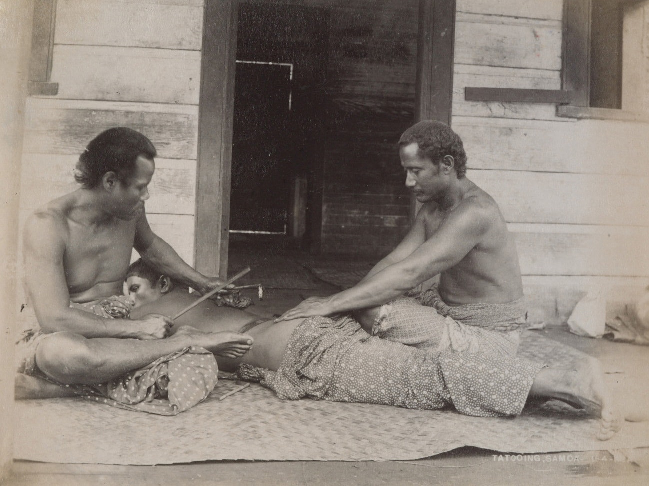 A sepia photo of two Sāmoan men tattooing another man who is lying down on a flax mat