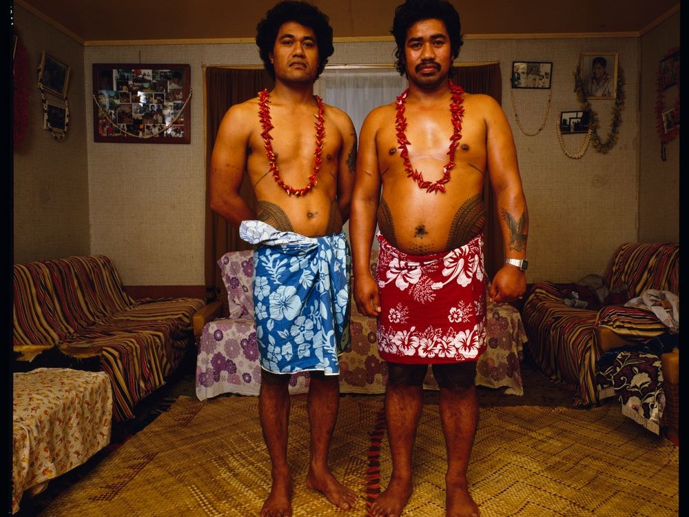 'Paul's nephew Jim at Paul's house', photo by Mark Adams, 1985. Te Papa (B.026409) Two Sāmoan men in lavalava's standing in a living room looking at the camera