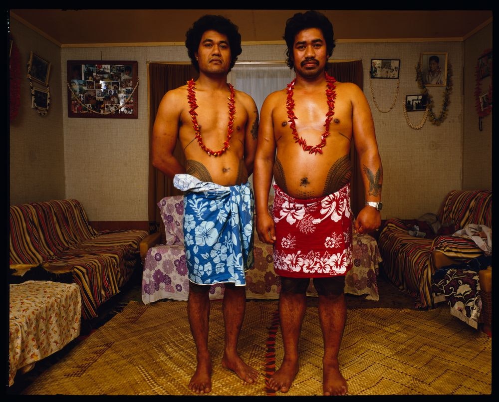 Two Sāmoan men in lavalava's standing in a living room looking at the camera