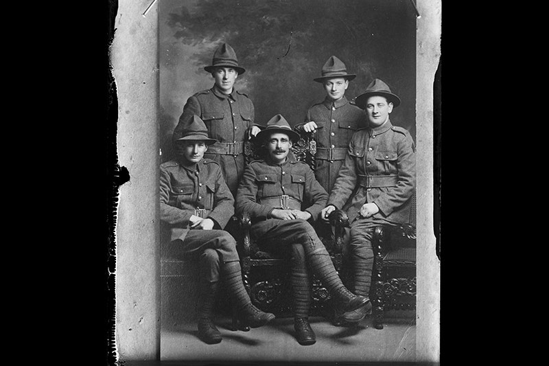 Five soldiers in a photographic studio. Three are sitting in a chair the others are standing.