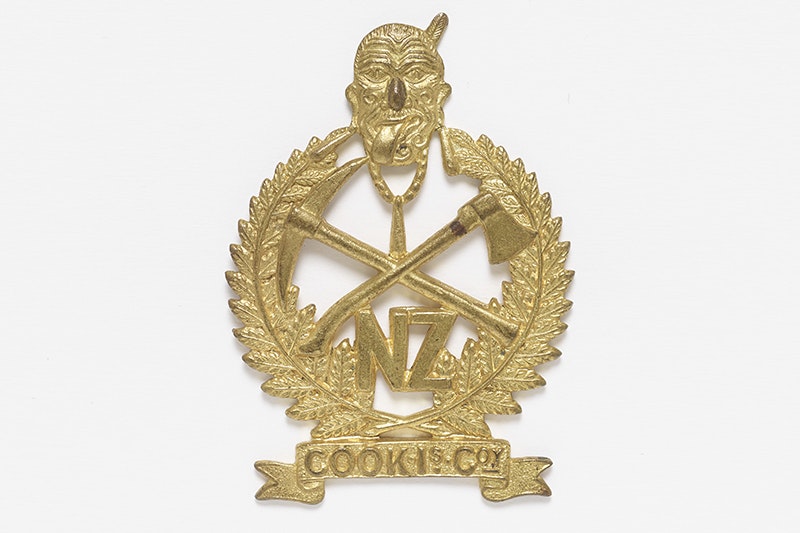 New Zealand Pioneer Battalion Badge and features the face of a Maori warrior wearing a solid necklace that joins the tips of two ferns. Crossed over in the centre is an axe and a pickaxe. "NZ" (an abbreviation of New Zealand) appears below the warrior’s n