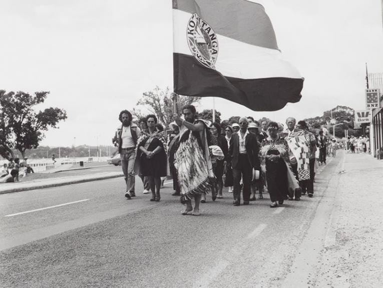 Black and white photo of people marching down a road. The person in front is holding a large flag