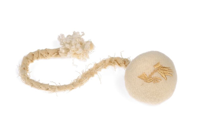 A round ball wrapped in a blanket material and a plaited cord attached. The ball has two gold hands on it with the wrists in chains.