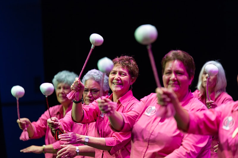Women all wearing black skirts and pink shirts swinging poi in unison (white balls on cord).