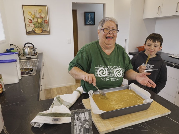 Still from Matariki kai: ginger crunch recipe, 2023. Te Papa Screenshot from a video, showing an older woman in a green T-shirt icing a cake in her kitchen as a young boy looks on