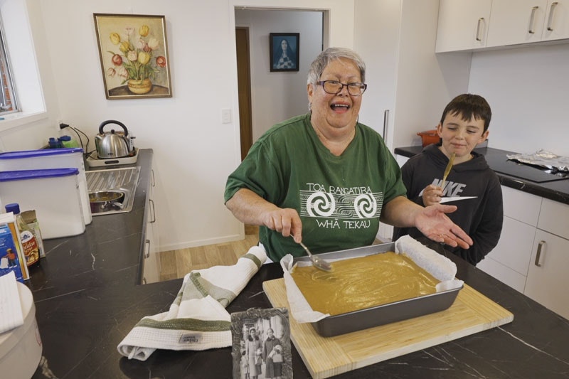 Screenshot from a video, showing an older woman in a green T-shirt icing a cake in her kitchen as a young boy looks on