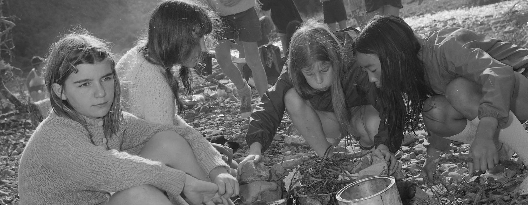 A group of young girls sitting by a river blow on twigs to ignite a fire