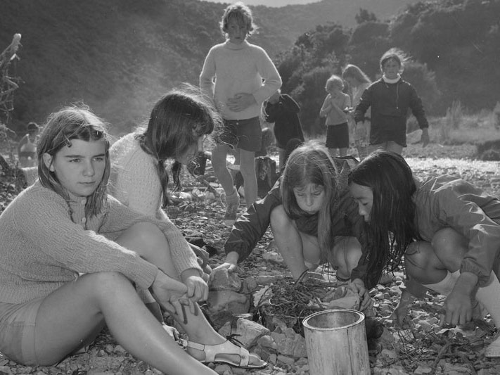 School camp activities – inscribed ‘Woolf (Camp)’, 1973–1975, Spencer Digby Studios, Wellington, black and white film negative. Spencer Digby / Ronald D Woolf Collection. Gift of Ronald Woolf, 1975. Te Papa (F.074184) A group of young girls sitting by a river blow on twigs to ignite a fire