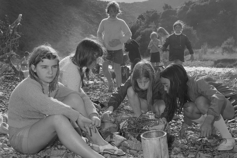 A group of young girls sitting by a river blow on twigs to ignite a fire