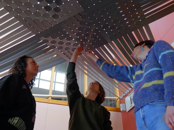Still from Making Takapau: Mataaho Collective discuss their monumental installation, 2023. Te Papa Still from a video. Shows three women underneath a large-scale weaving that they are working on, looking at its finer details