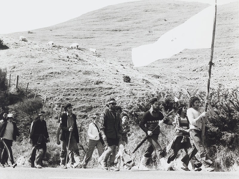 Māori Land March, 1975, by Ans Westra, gelatin silver print. Purchased 1993 with New Zealand Lottery Grants Board funds. Te Papa (O.009298/03) Black and white photo of a large group of people marching alongside a rural road. The person at the front is carrying a large flag