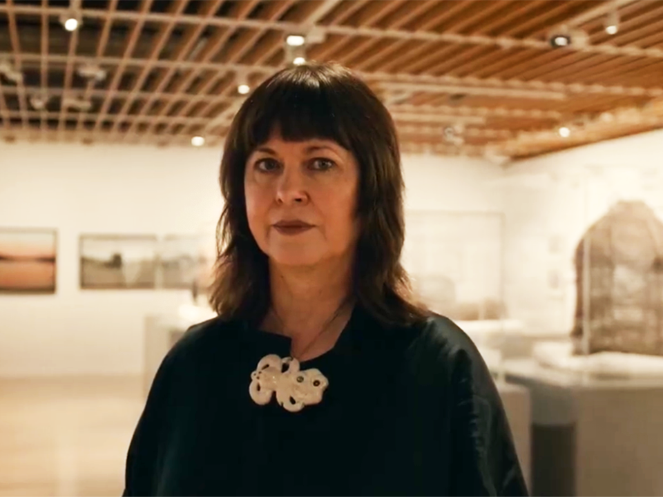 Still from Natalie Robertson video. Te Papa Head and shoulders of a woman with dark hair and dressed in black standing in a gallery