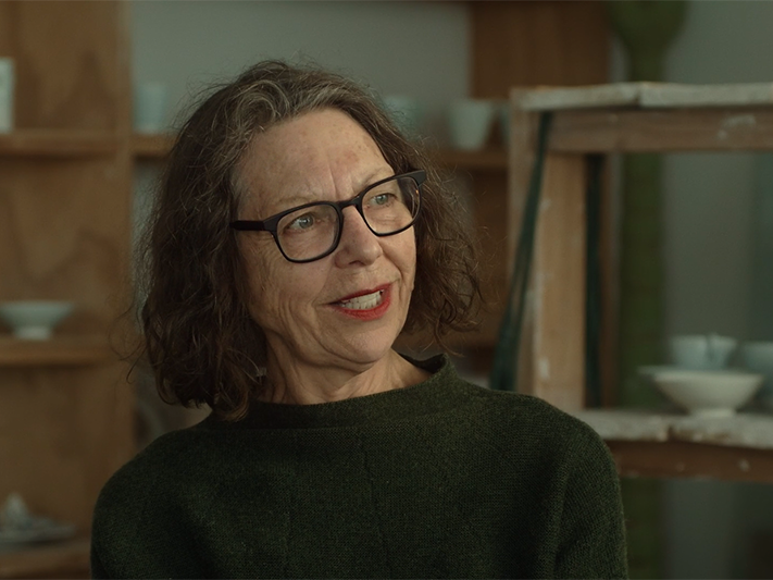 Still from video of Raewyn Atkinson in her studio A woman talking to someone off screen and sitting in an artist studio.