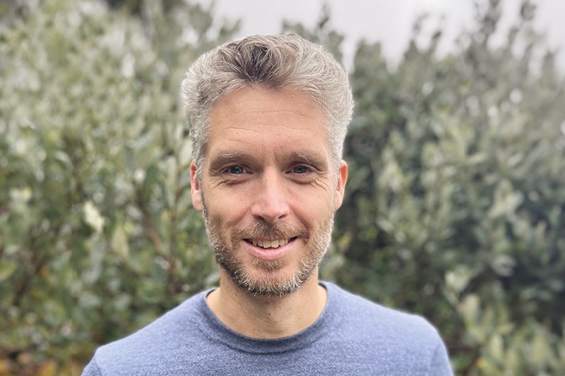 Head and shoulders of a white man with grey hair and a blue jumper. He is standing in front of green shrubbery.