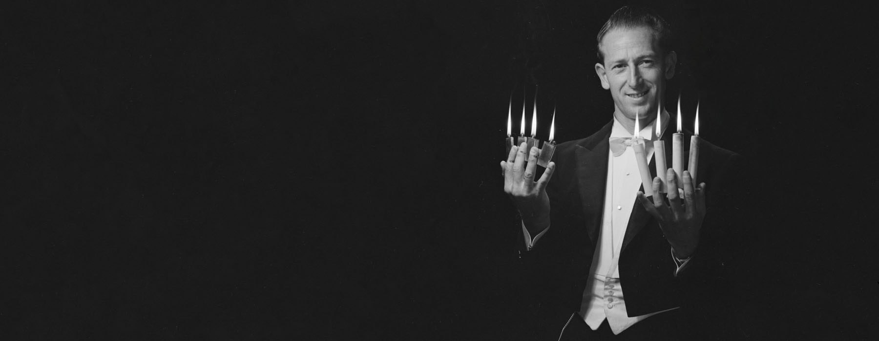 A magician poising for a photo. In his left hand area lit cigarette lighters between each finger. In his right handle, lit candles are held between each finger