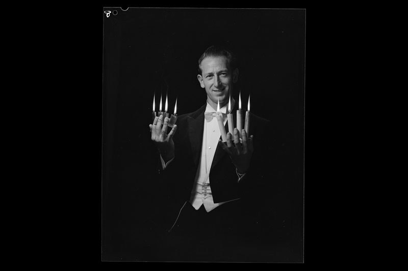 A magician poising for a photo. In his left hand area lit cigarette lighters between each finger. In his right handle, lit candles are held between each finger