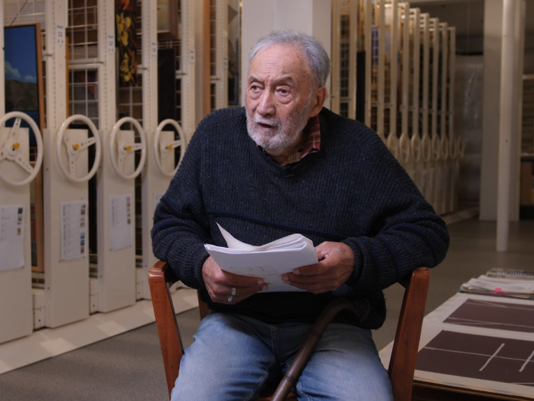 An older man sitting in a chair holding A4 sheets of paper and talking to someone off camera. There are rolling files behind him.