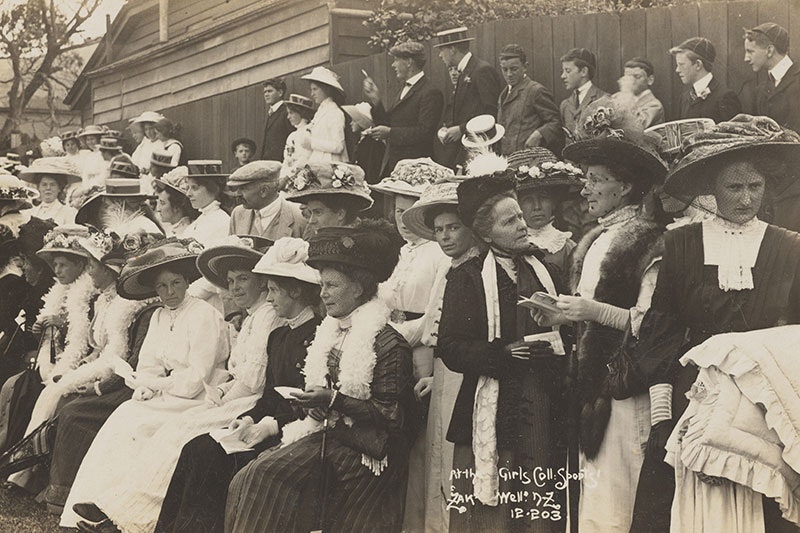 A large group of women sitting watching something out of frame. Men stand behind them