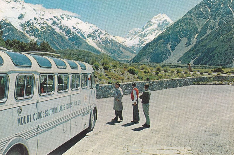 View of Mount Cook from a carpark. A bus can be seen to the left and three people on the right stand looking at the mountain
