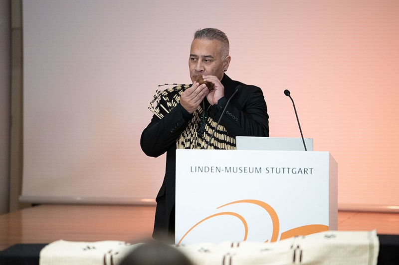 A man is standing at a podium, playing a small indigenous flute into a microphone