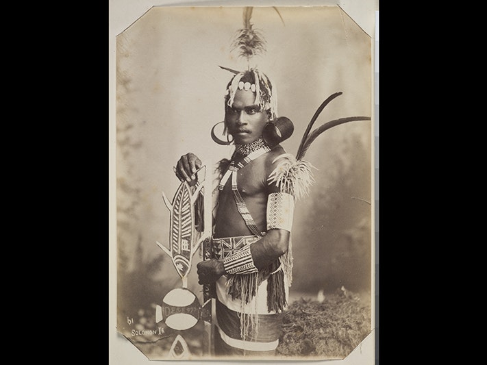 Unidentified Solomon Island Man, 1890-1910, by Thomas Andrew. Te Papa (O.002242) Studio portrait of a young man standing, 3/4 profile view. He is wearing finely beaded strings across his chest, upper arms and forearm. He is also wearing a beaded waistband which has a British flag on it. In addition he has two large, cylindrical earrin
