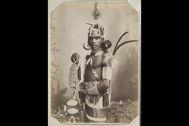 Studio portrait of a young man standing, 3/4 profile view. He is wearing finely beaded strings across his chest, upper arms and forearm. He is also wearing a beaded waistband which has a British flag on it. In addition he has two large, cylindrical earrin