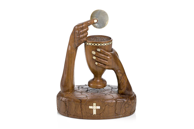 A carved wooden sculpture of two hands - one hand is holding a chalice and the other hand is holding a wafer over the chalice.