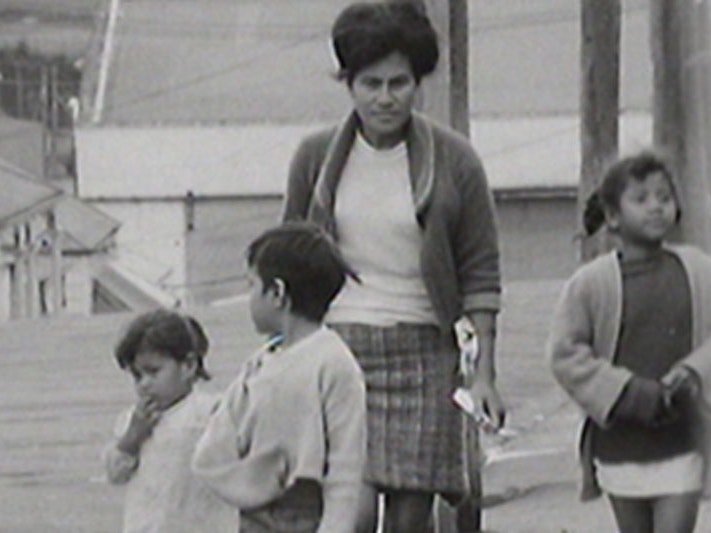 Still from Dawn Raids. NZ On Screen Black and white still from a video, showing a Polynesian women and three children walking down a street
