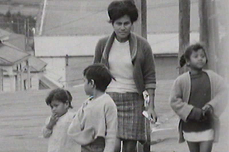 Black and white still from a video, showing a Polynesian women and three children walking down a street