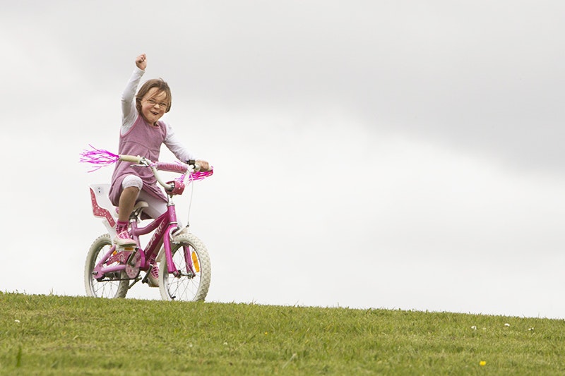 A child in a pink and white top riding a bright pink pushbike with pink tassells over grass and is punching the air with her right fist.
