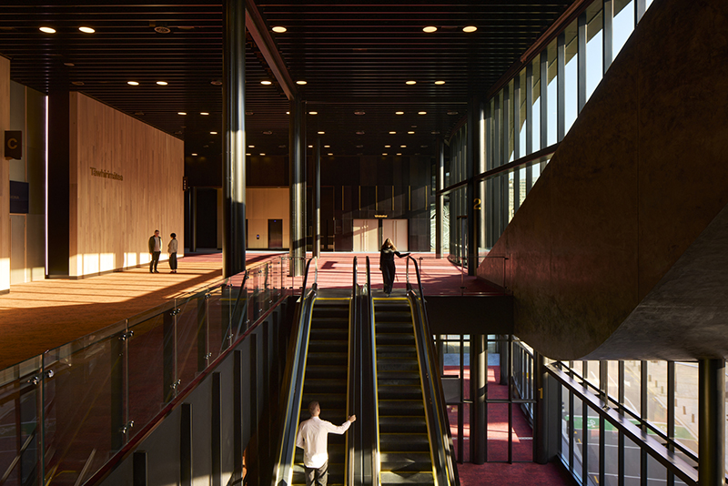 Inside a convention centre with escalators and people walking around.