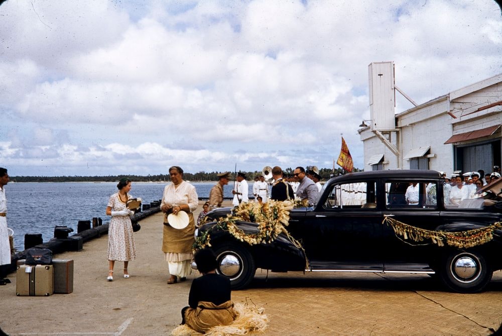 A photo of the British Queen and the Tongan queen walking on a waterfront. There is a ceremonial car parked nearby.