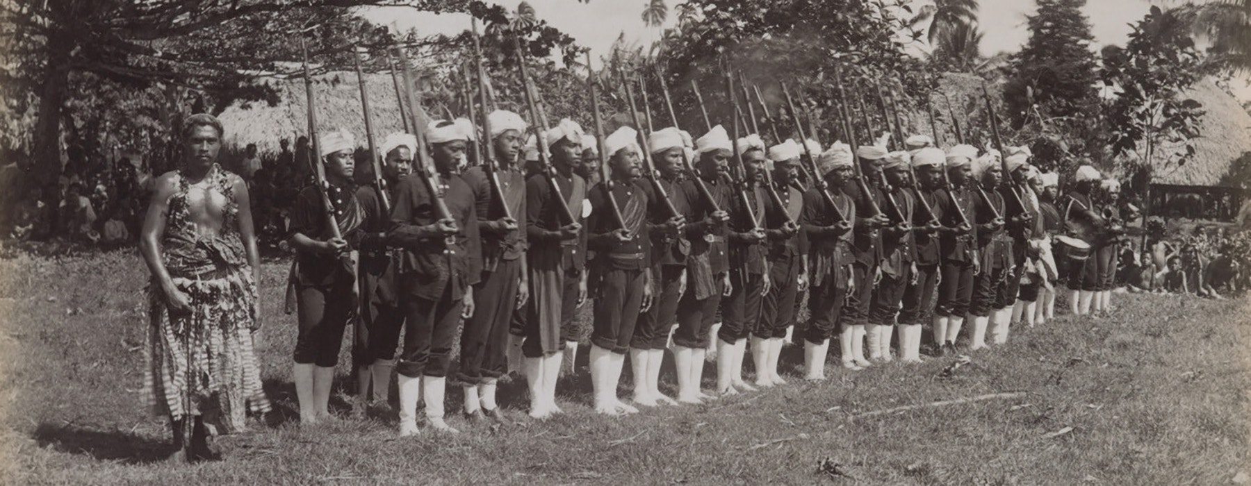 A sepia photo of a contingent of soldiers with white boots and rifles and white headwear. There is one person at one end in Sāmoan traditional costume