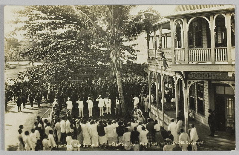 A sepia photo of people in uniform in Sāmoa hoisting a flag - they are surrounded by troops and onlookers.