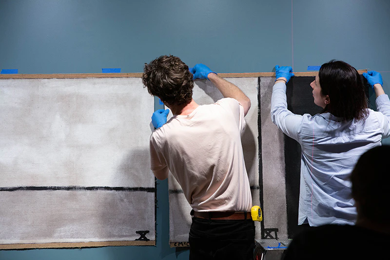 Two people with their backs to the camera and wearing blue gloves are hanging a large painting on the wall.