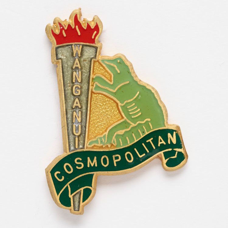 Decorative badge featuring a large flaming torch with the word Wanganui on it, and a green lizard standing beside it looking upwards at the flame. Underneath appears the word ‘cosmopolitan’