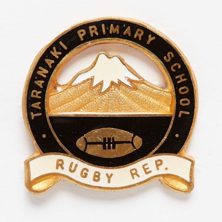 Black and gold circle-shaped badge featuring depiction of Mt Taranaki with a rugby ball underneath it and a sash with the words ‘Taranaki Primary School Rugby Rep’