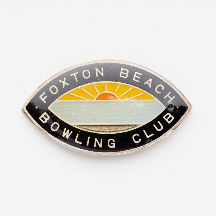 Eye-shaped badge with a black border featuring the words ‘Foxton Beach Bowling Club’. Inside the border is a colourful depiction of a sunset from a beach
