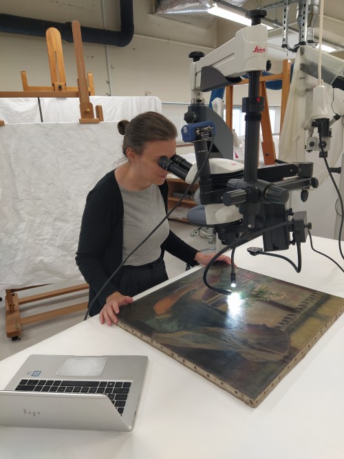 A woman looks at an oil painting through a large microscope.