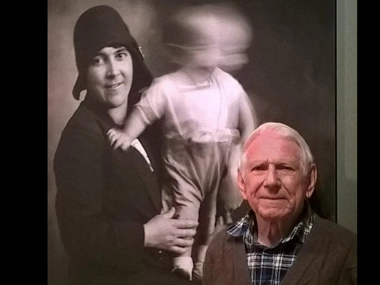 An elderly man is standing in front of a blown-up photo of a woman holding a baby. The baby is blurry because he is moving is head.