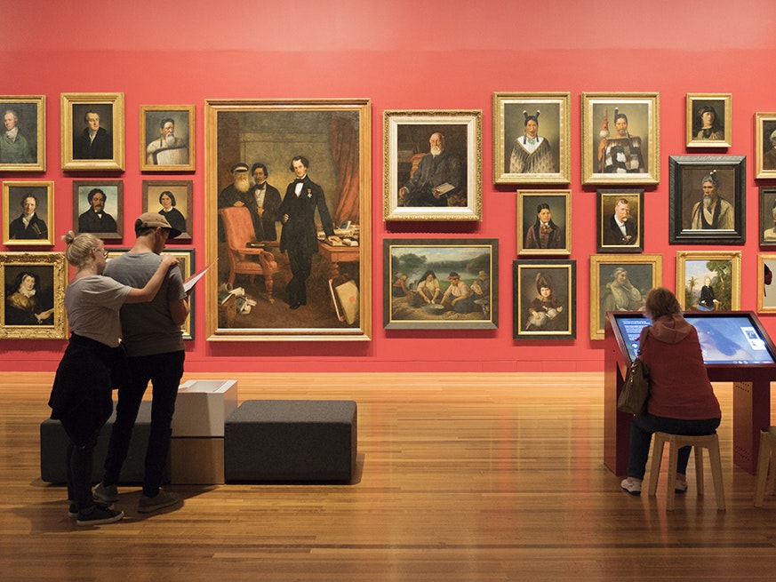 Toi Art Portrait Wall Digital Labels interaction, 2018. Photograph by Kate Whitley. Te Papa (106646) People in a gallery looking at paintings on the wall. One person is sitting in front of a large screen.