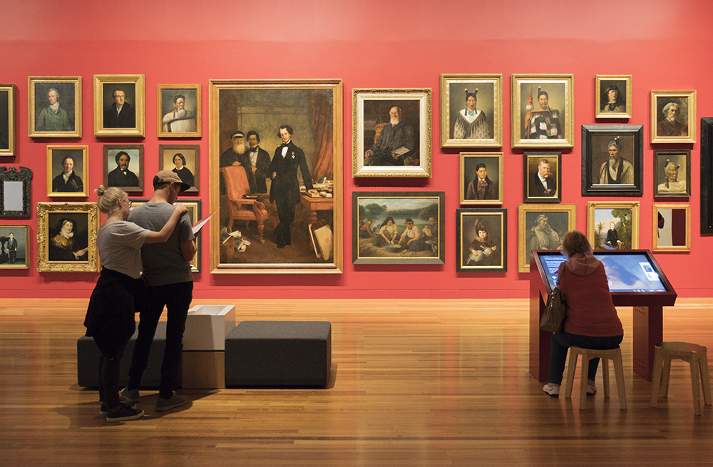 People in a gallery looking at paintings on the wall. One person is sitting in front of a large screen.