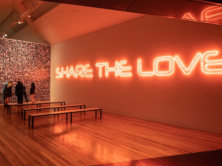 Visitors looking at the installation Top16, 2007-2017 by Janet Lilo, 2019. Photo by Johnny Hendrikus. Te Papa (136763) People in a large room that has a giant neon sign that says Share the love.