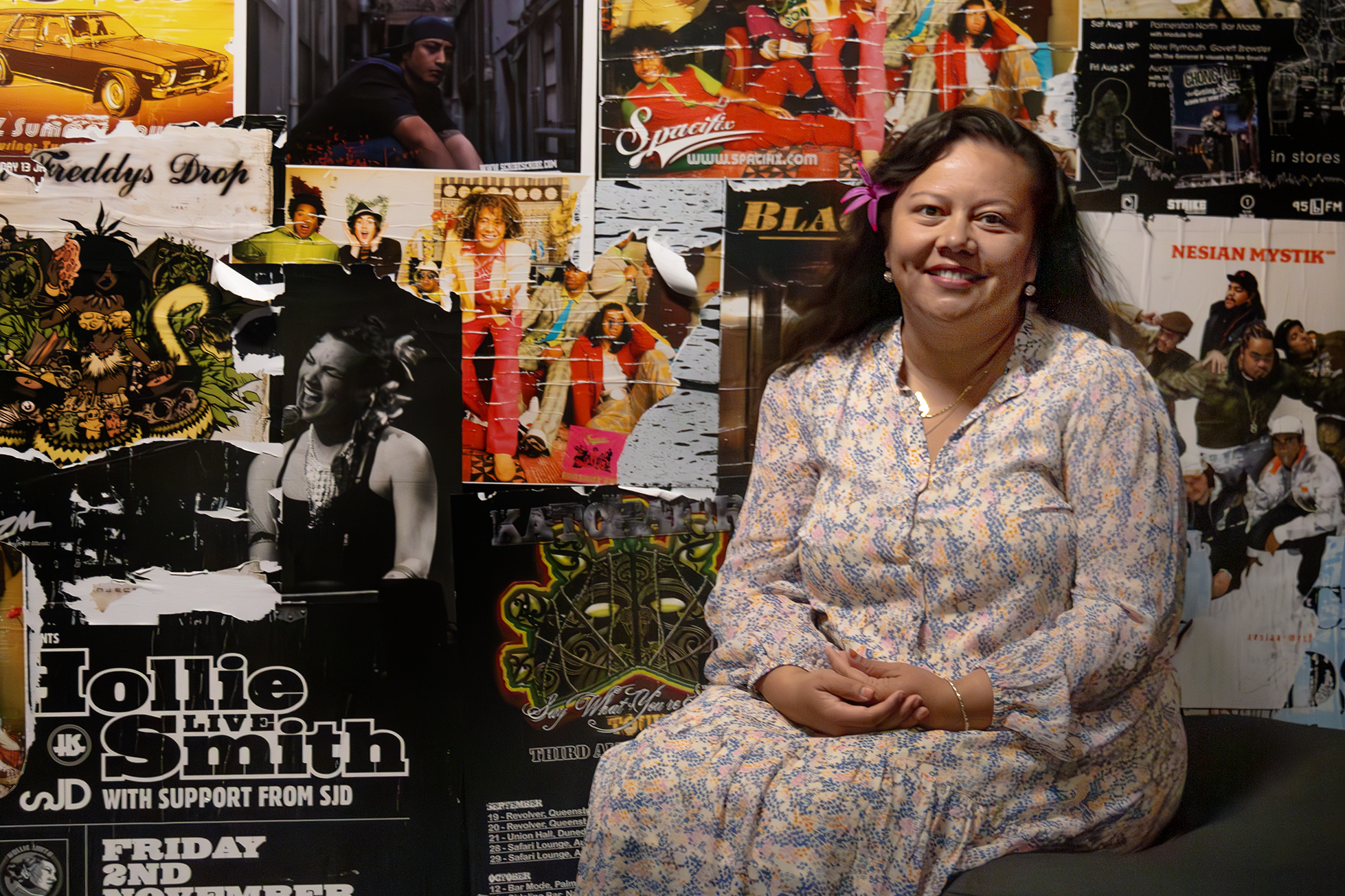 A woman in a light-coloured dress is sitting in front of a wall of music posters. She is posed and smiling at the camera.